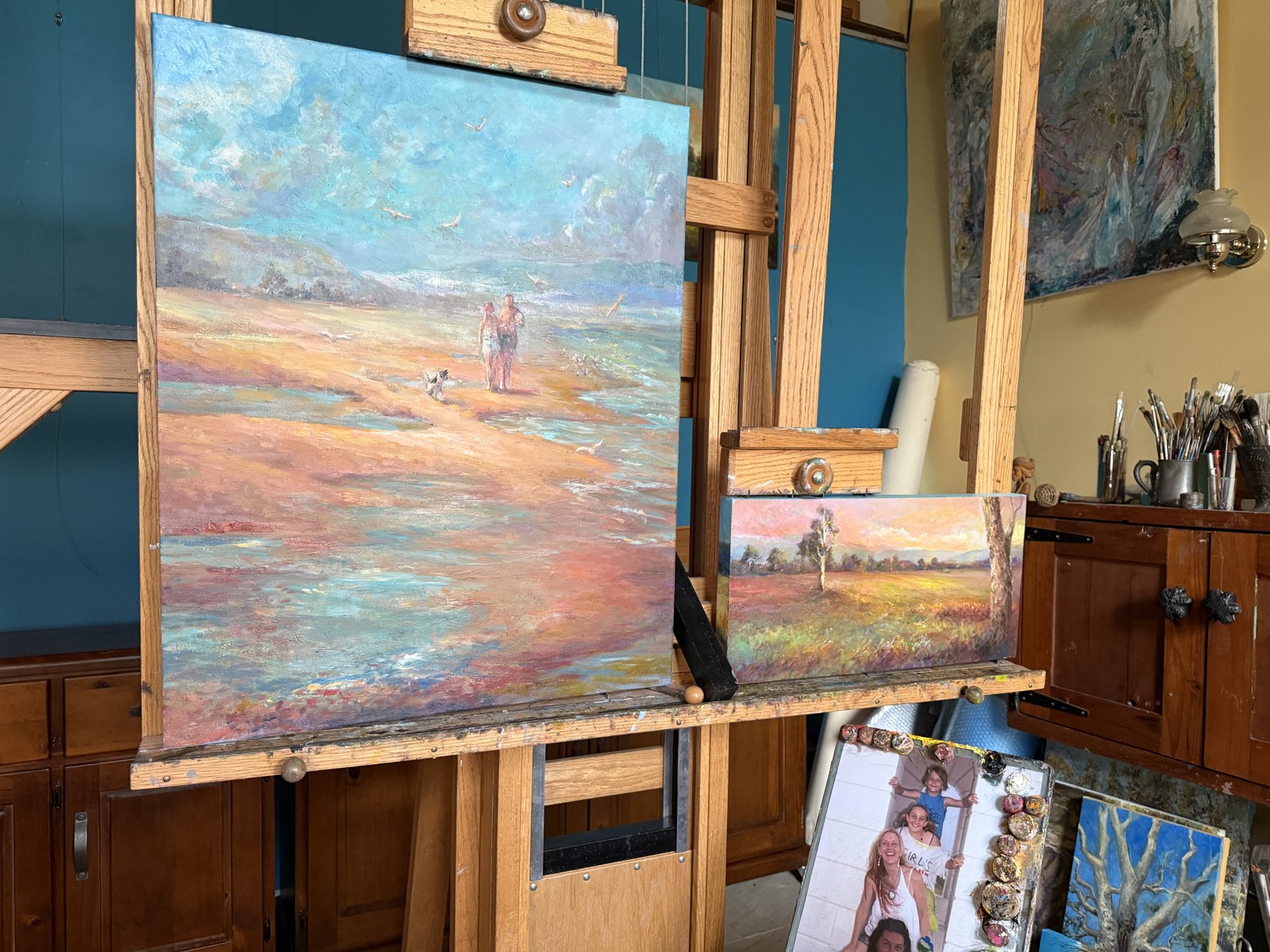 The two paintings together on the easel, a beach scene with figures and a small golden landscape with a lone tree.